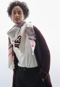 Écharpe multicolore avec un bord frangé, motif à carreaux dans des tons de blanc, gris et rose, avec un texte en gras de couleur rouge foncé.