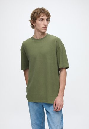 Junger Mann mit lockigem Haar, der ein olivgrünes Kurzarmhemd und hellblaue Jeans trägt und nach links blickt vor einfachem Hintergrund.