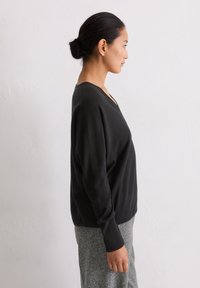 Schwarzer Wollpullover mit tiefem V-Ausschnitt, fallenden Schultern und langen Ärmeln, kombiniert mit grauem Material darunter, glatte Textur.