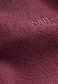 Bordeaux puuvillakangas, jossa on pehmeä tekstuuri ja jonka pinnalla on hienovarainen brodeerattu logo, joka lukee "self." vaaleammalla langalla.