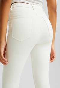 Närbild av en person i högmidjade vita jeans med bakfickor och en ribbad vit topp, med avslappnade händer vid sidorna.