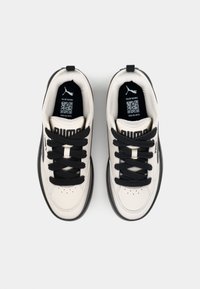 Tênis brancos e pretos com um cabedal liso, cadarços pretos grossos, colarinho acolchoado, alça fácil para puxar e perfurações visíveis na caixa dos dedos.