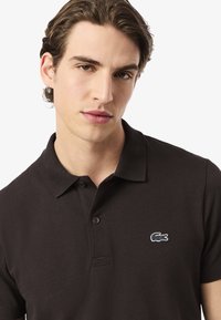 Ung man med vågigt brunt hår som bär en svart pikétröja med en liten krokodil-logotyp på vänster bröst, mot en enkel ljus bakgrund.