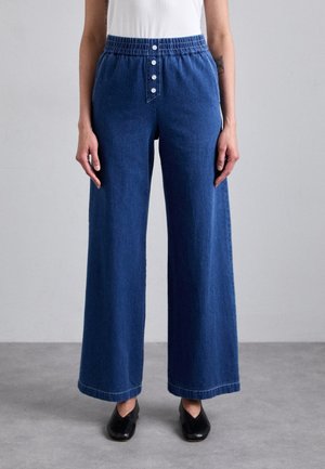 Persoon draagt blauwe wijde denimbroek met elastische taille en knoopdetail aan de voorkant, gecombineerd met zwarte schoenen en een wit topje.