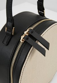 Sac à main rond avec dessus en cuir noir, devant tissé beige, fermeture éclair dorée, et tirettes de fermeture éclair ainsi que anses en cuir noir.