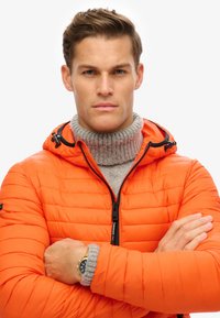 Pufjakke i lys orange med quiltet tekstur, lynlåset front og hætte. Stylet med en grå turtleneck under.