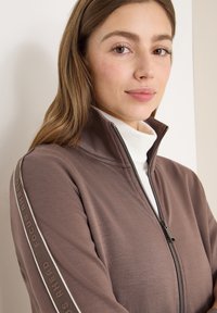 Hnědá bunda na zip s vysokým límcem, s bílým zipem a bočním páskem. Text "FOCUS AHEAD" potištěný podél rukávů. Měkká textura.