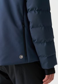 Námořnická puffer bunda s prošívanou texturou, která má zipovou kapsu a detail v podobě kruhového loga blízko lemu. Měkký, matný povrch.