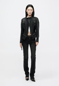 Μαύρο κοντό δερμάτινο μπουφάν με δύο μπροστινές τσέπες, σε συνδυασμό με ψηλόμεσα μαύρα τζιν και μαύρες μυτερές γόβες, σε λευκό φόντο.