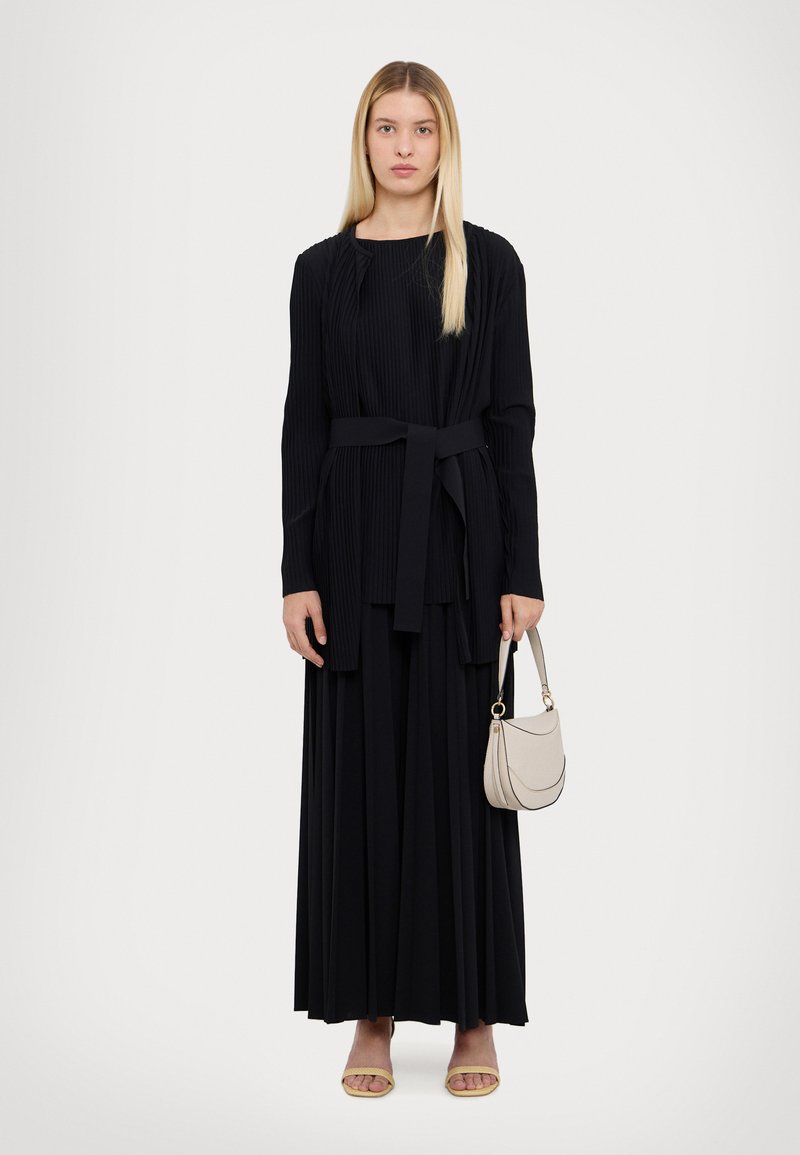 Schwarzes, geflämmtes Maxikleid mit einem gebundenen Taillengürtel, kombiniert mit einer kleinen beigen Handtasche und flachen Sandalen. Einfacher, runder Ausschnitt und lange Ärmel.