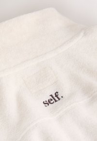 Tissu polaire écru avec un patch cousu et le mot « self. » brodé en fil foncé.