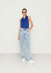 Top halter azul, jeans de cintura alta azul claro e ténis brancos. Óculos de sol escuros e brincos de argola acrescentam acentos distintos.