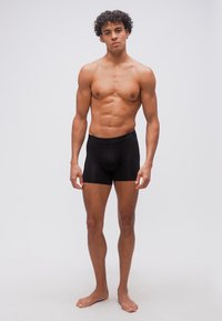 Νεαρός άνδρας με σγουρά μαλλιά, χωρίς μπλούζα, όρθιος, ξυπόλυτος, φορώντας μαύρο μπλεκάκι, σε απλό λευκό φόντο.