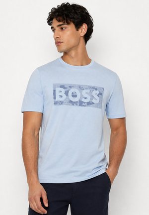 Homme aux cheveux bouclés et foncés portant un T-shirt bleu clair avec le texte « BOSS » en motif camouflage, associé à un pantalon foncé, debout sur fond blanc.