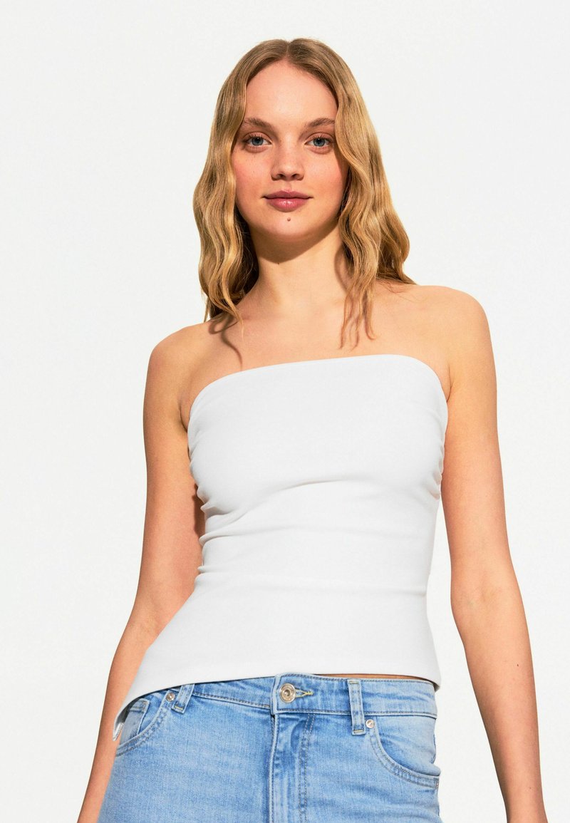 Top bianco senza spalline con un design aderente, realizzato in tessuto morbido, abbinato a jeans di denim azzurro chiaro, che mostrano un'estetica pulita e semplice.