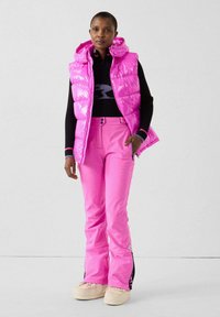 Gilet imbottito rosa chiaro con cappuccio, abbinato a pantaloni da sci rosa coordinati. Maglia nera a maniche lunghe con logo. Tessuto morbido e lucido con design sportivo.