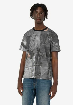 Młody mężczyzna z krótkimi dredami, ubrany w czarno-biały patchworkowy t-shirt z nadrukiem dżinsowym i niebieskie dżinsy, stoi na białym tle.