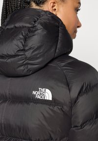 Casaco puffer preto com um colarinho alto, apresentando um logótipo branco do lado esquerdo. Exterior suave e brilhante com costuras acolchoadas.
