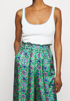 Kvinde iført en hvid tanktop med blå og grøn kant og højtaljede grønne bukser med et farverigt blomsterprint og lommer.