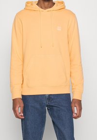 Sweat à capuche jaune en coton avec une poche avant, une capuche à cordon de serrage et un petit patch logo. Associé à un jean en denim bleu foncé.