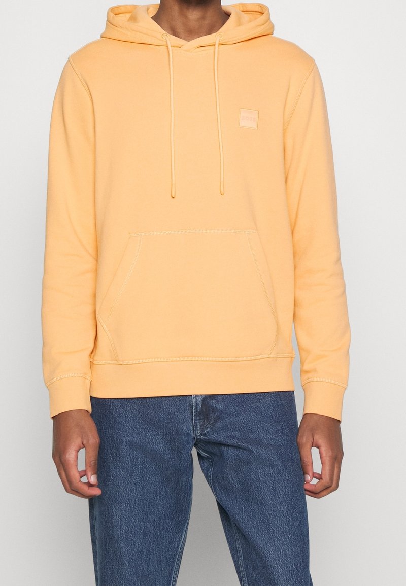 Sweat à capuche jaune en coton avec une poche avant, une capuche à cordon de serrage et un petit patch logo. Associé à un jean en denim bleu foncé.