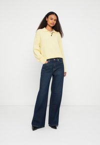 Yellow collared sweater with three buttons, paired with dark blue wide-leg jeans and black heeled shoes. Model stands with one hand in pocket.