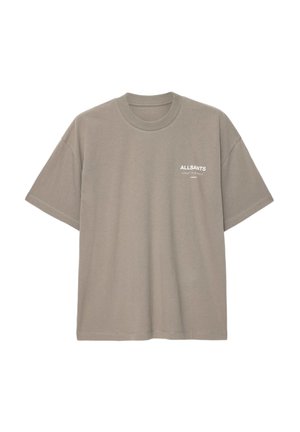 Personne aux cheveux tressés portant un t-shirt oversize taupe, un pantalon blanc et des baskets noires, tenant un sac cabas noir à ses côtés.