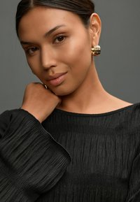 Schwarze, strukturierte Bluse mit weiten, gerüschten Ärmeln. Goldene Ohrstecker im linken Ohr. Glatte Hautfarbe, weiche Gesichtszüge und schlichter Hintergrund.