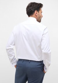 Weißes Langarmhemd mit einem hebbaren Kragen, kombiniert mit dunkelblauen Hosen, die seitliche Taschen und Manschetten mit individuellen Hardware-Akzenten aufweisen.