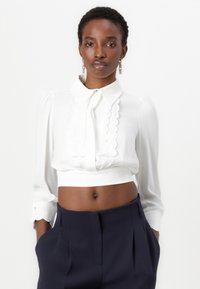 Blusa bianca corta con colletto scallop e maniche lunghe, caratterizzata da una vita arricciata e dettagli con bottoni ai polsini.