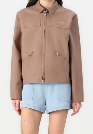 Bruine jas met ritssluiting, kraag en diagonale zakken met rits, gedragen over lichtblauwe denimshorts bij een staand persoon.
