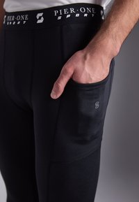 Schwarze Sporthose mit weichem Material, ausgestattet mit einem logoartigen Bund, einer Seitentasche und verstärkten Nähten für zusätzliche Haltbarkeit.