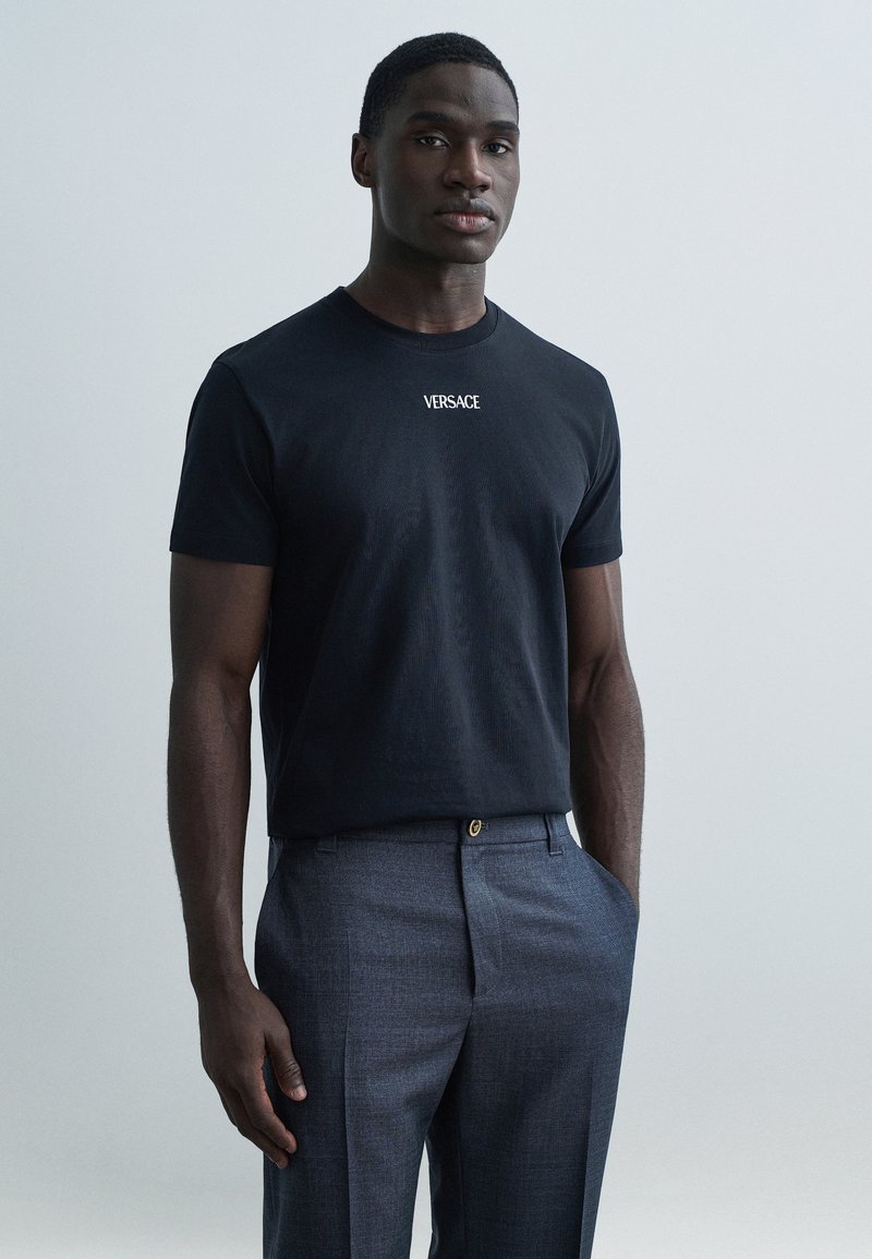 Μαύρο βαμβακερό T-shirt με το λογότυπο «VERSACE» σε λευκό, συνδυασμένο με γκρι υφασμάτινο παντελόνι, που παρουσιάζει μοντέρνα γραμμή και κομψό σχεδιασμό.