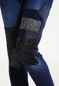 Jeans de mezclilla azul oscuro que presentan varios segmentos de patchwork en telas negras y grises, con acentos texturizados y un diseño ajustado alrededor de las rodillas.