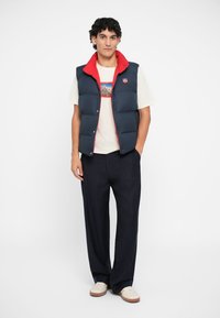 Navy sleeveless puffer vest with red accents, worn over a cream t-shirt featuring a mountain graphic, paired with pinstriped dark pants.