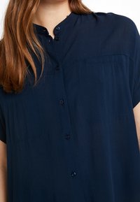 Camicia a maniche corte blu navy con un design a bottoni, tessuto morbido, colletto rotondo e due taschini sul petto. Presenta una vestibilità comoda.