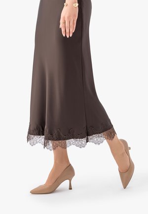 Kobieta w brązowej spódnicy z koronkowym wykończeniem i beżowych szpiczastych butach na niskim obcasie, z złotą bransoletką i pierścionkiem na lewej ręce.