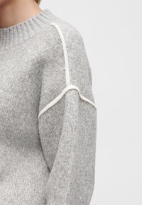 Gros plan d'une personne portant un pull en maille gris clair avec des détails de coutures blanches en relief le long de l'épaule et de la manche.
