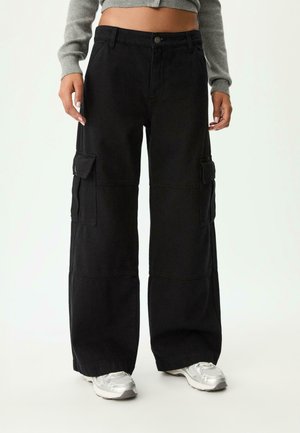 Osoba w czarnych cargo spodniach o szerokich nogawkach i wysokim stanie z kieszeniami z klapami, szarym krótkim swetrze oraz białych masywnych sneakersach stoi z luźno opuszczonymi rękami.