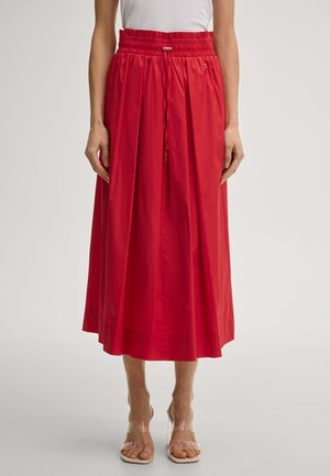 Vrouw draagt een rode midi-rok met hoge taille en plooien, een trekkoord in de taille en doorzichtige sandalen met hak, staand tegen een effen achtergrond.