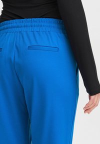 Närbild av en person som bär ljusblå byxor med elastisk midja och fickor med kant, tillsammans med en figurnära svart långärmad topp.