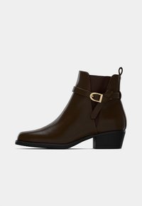 Botín de cuero marrón oscuro con tacón bajo, panel elástico lateral, correa decorativa y hebilla en tono dorado sobre fondo blanco.