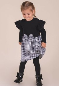 Czarna sukienka z długim rękawem, z falbankami na ramionach i spódnicą w pepitkę, ozdobiona kokardą, zestawiona z czarnymi legginsami i butami.