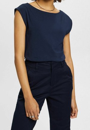 Osoba ubrana w granatowy top bez rękawów i pasujące spodnie z wysokim stanem, z łańcuszkowym naszyjnikiem oraz pierścionkami na lewej ręce.