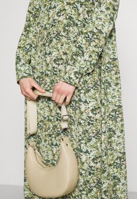 Beige Lederhandtasche mit geschwungener Form und einem abnehmbaren beigen Tragegurt, in der Hand gehalten, neben einem grün gemusterten Kleid mit Blumen.