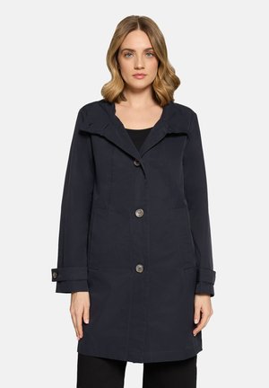 Frau trägt einen schwarzen Mantel mittlerer Länge mit großen Revers, Knopfverschluss, Seitentaschen und geknöpften Manschetten.