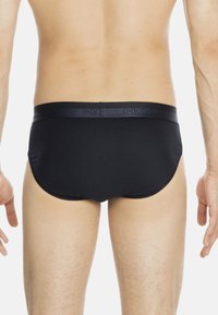 Mann in schwarzen Unterhosen, der mit dem Rücken zur Kamera steht, vor einem schlichten weißen Hintergrund.