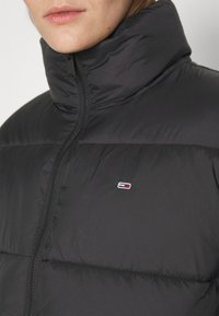 Giubbotto imbottito nero con colletto alto e chiusura con zip. Presenta una texture trapuntata e una piccola patch con logo in rosso, bianco e blu sul petto.