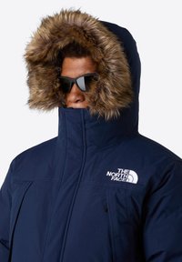 Parka blu navy con cappuccio foderato in pelliccia sintetica, alto colletto, chiusura con zip e logo The North Face ricamato sul petto.