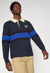 Pullover a maglia grigio scuro con colletto beige, caratterizzato da righe orizzontali blu e bordeaux e un logo sul petto. Maniche lunghe, vestibilità comoda.